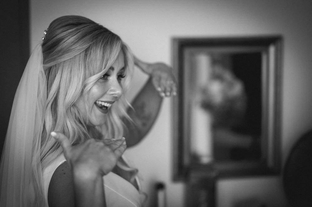 Black and white photo of a smiling bride in a veil adjusting her hair, with a mirror reflection in the background.