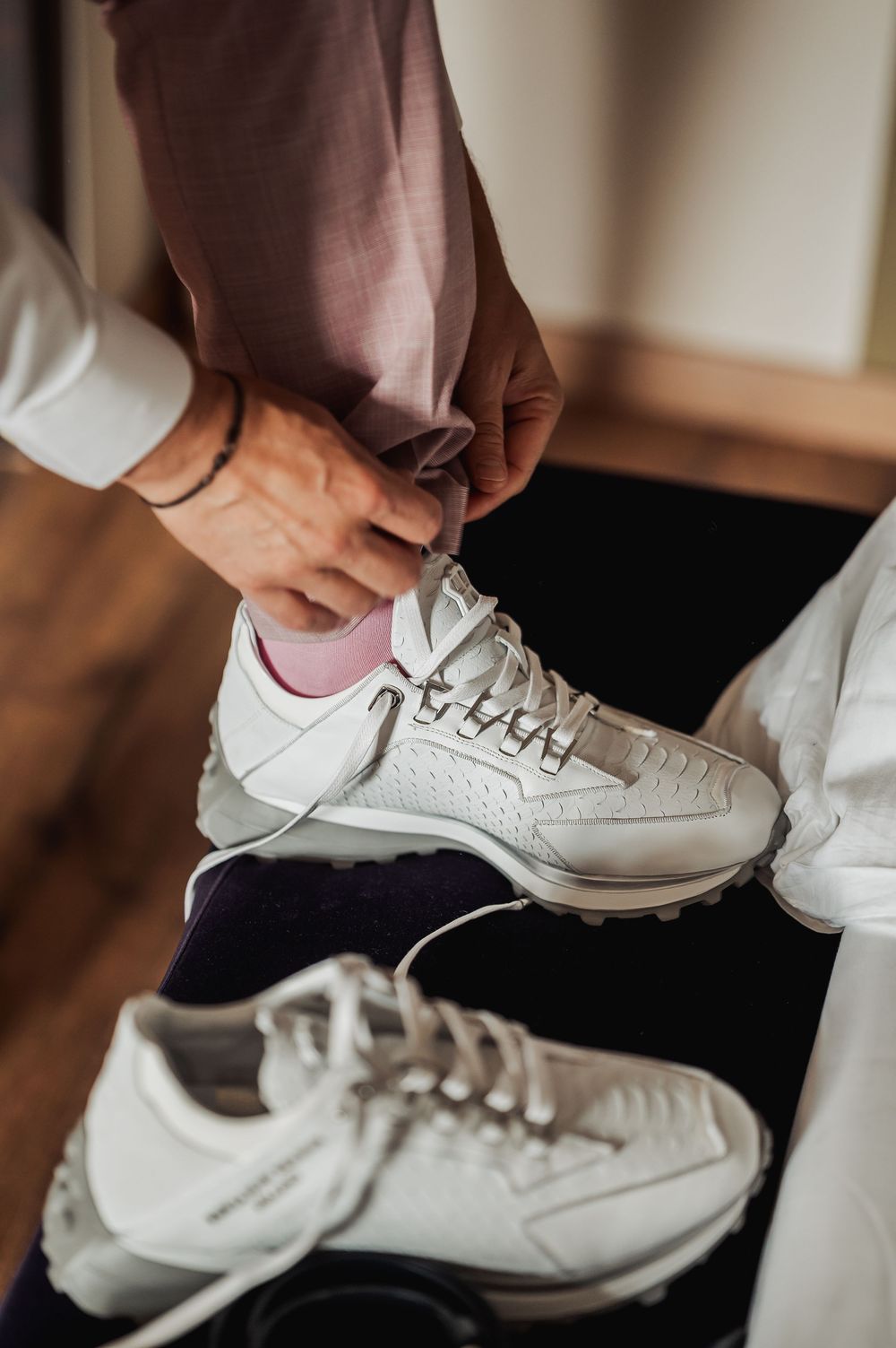 Person tying white sneakers while wearing pink socks and mauve trousers, with a second pair of white shoes on the floor.