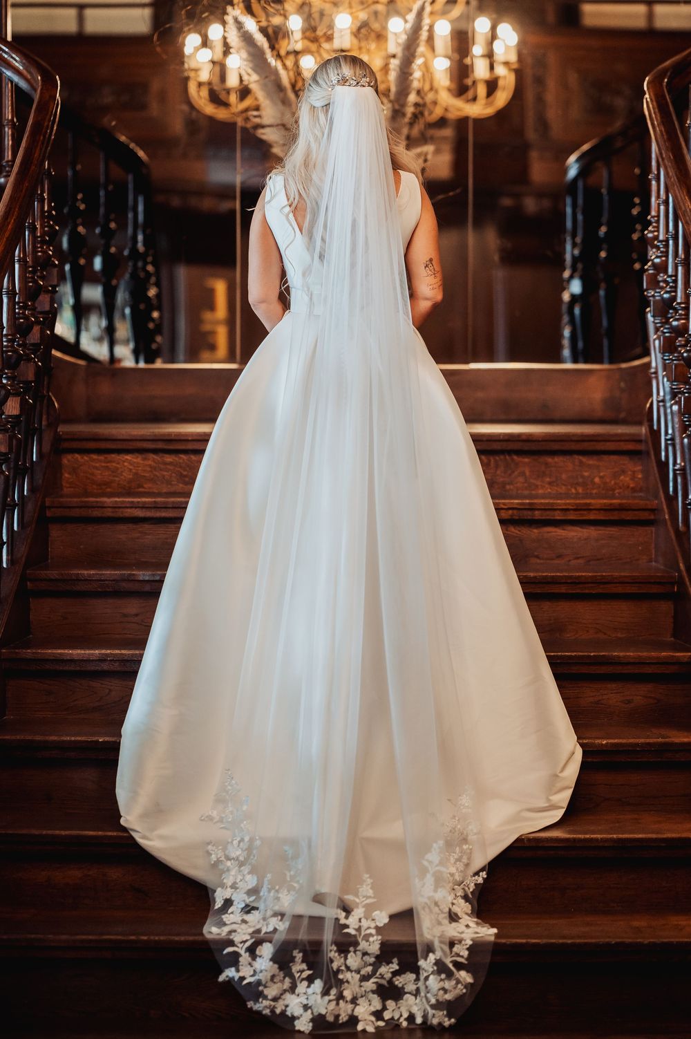 Bride in elegant white ball gown with lace-trimmed veil stands on wooden staircase in grand venue.