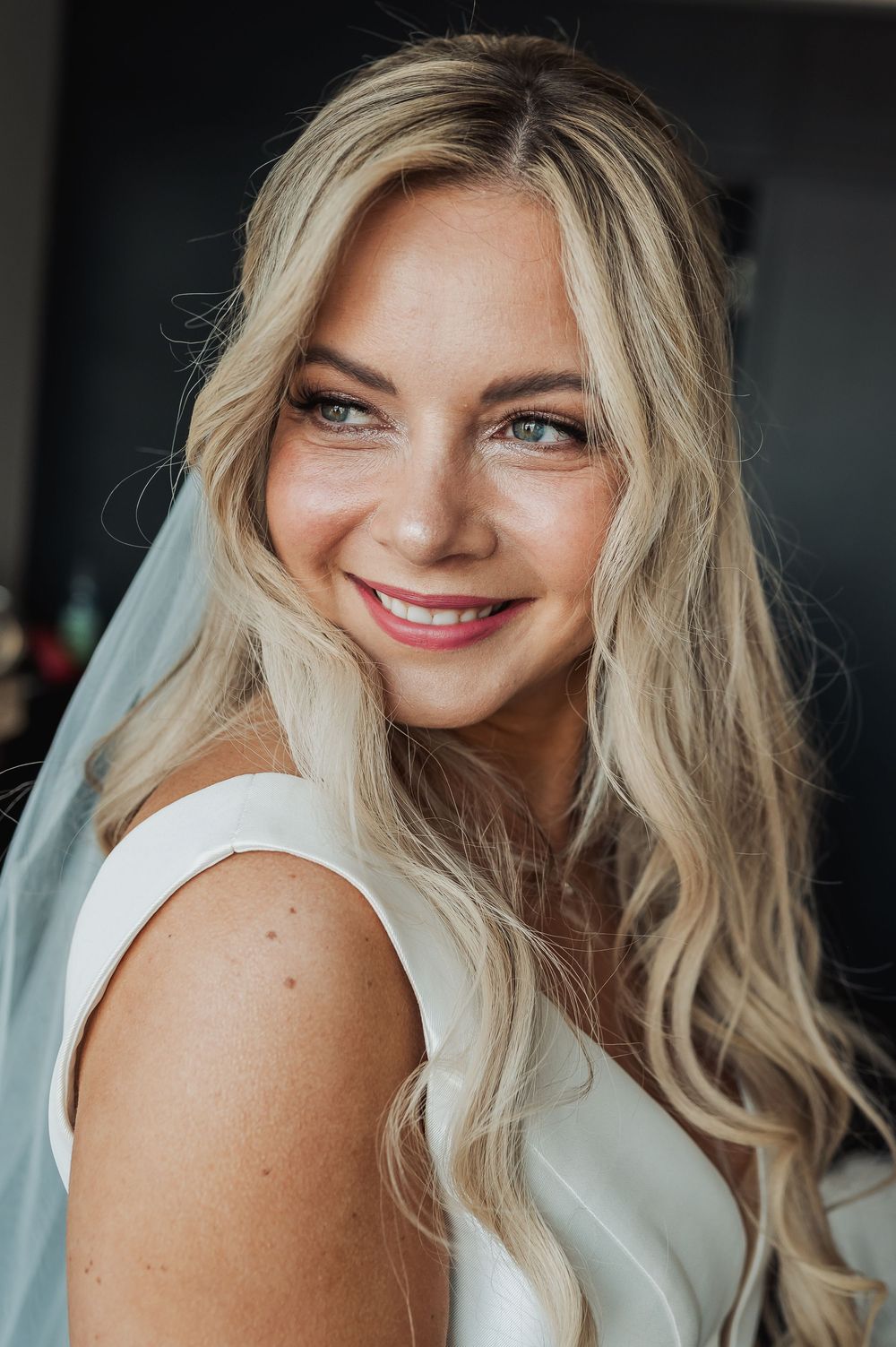 Smiling blonde bride with blue eyes wearing a white dress and veil, with wavy hair and pink lipstick.