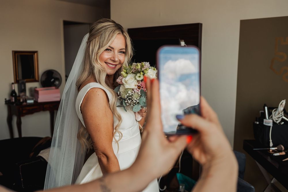 Bride in white gown and veil holding bouquet smiles as someone takes her photo on a smartphone indoors.