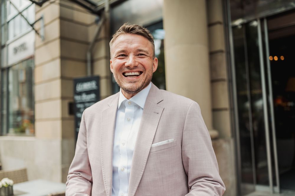 Smiling man in a pink blazer and light blue shirt standing outside a modern building.