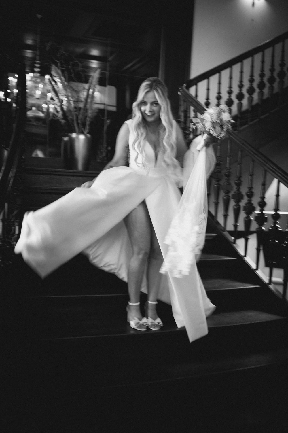 Bride in white gown holding bouquet descends grand staircase, black and white wedding photo.