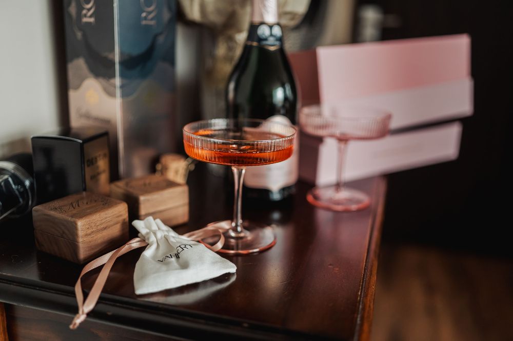 Elegant pink coupe cocktail glass on dark dresser with champagne bottle and wedding accessories.