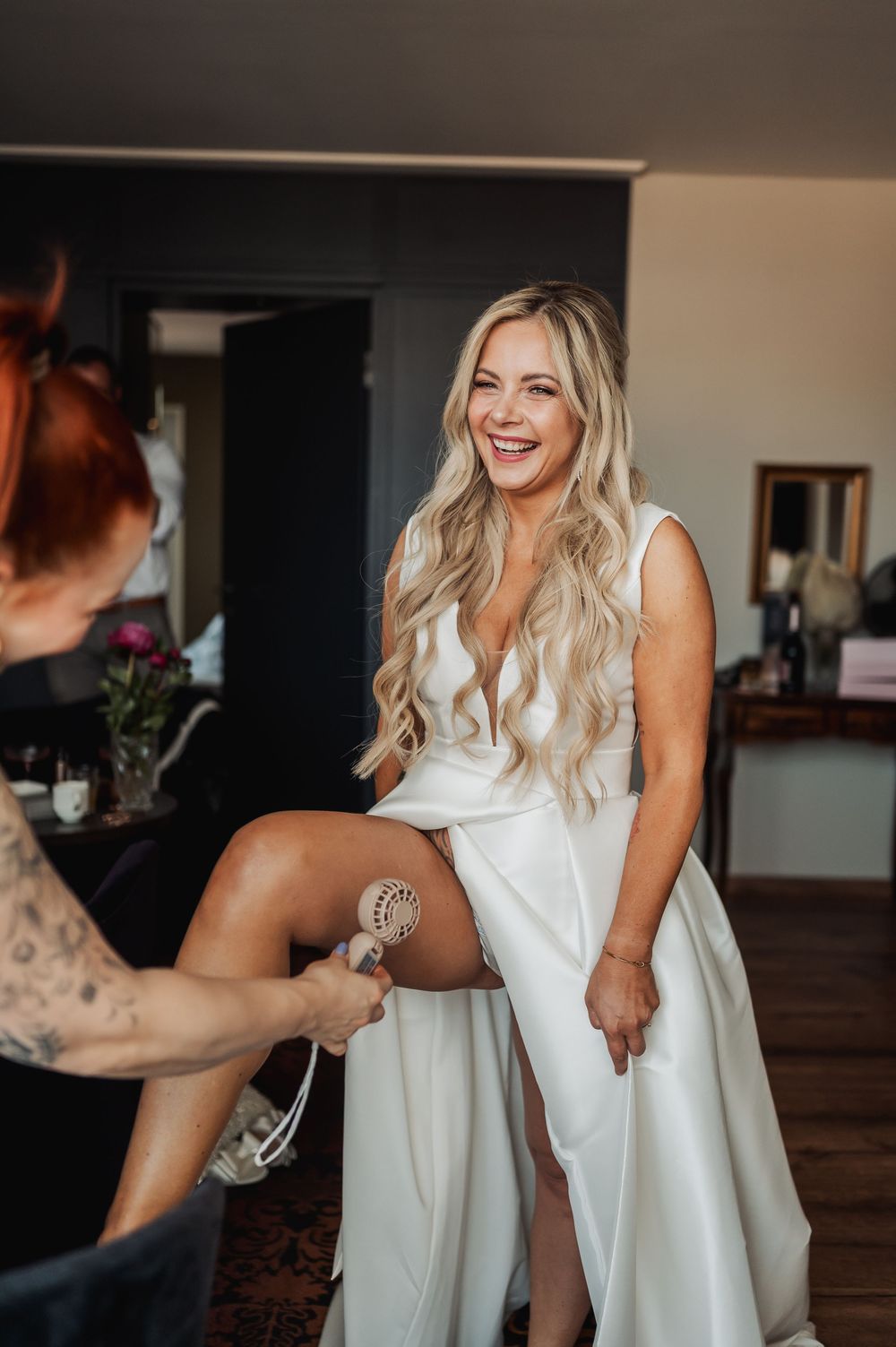 Laughing bride in sleeveless white gown gets bridal garter placed by bridesmaid on wedding day.