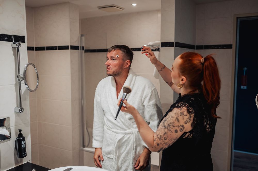 Woman with red hair applying makeup to man in white robe in modern hotel bathroom mirror.