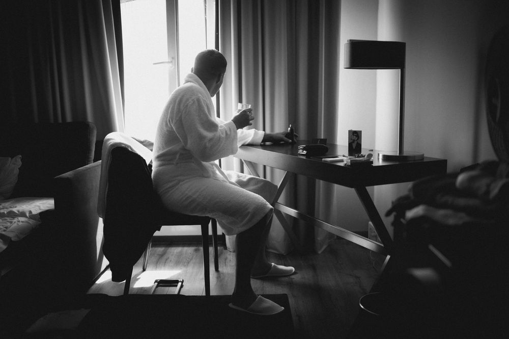 Black and white photo of groom in white suit sitting by window in hotel room before wedding.