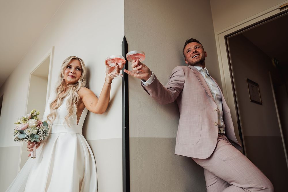 Bride and groom playfully toasting with pink drinks on their wedding day, bride holding bouquet in elegant white gown.