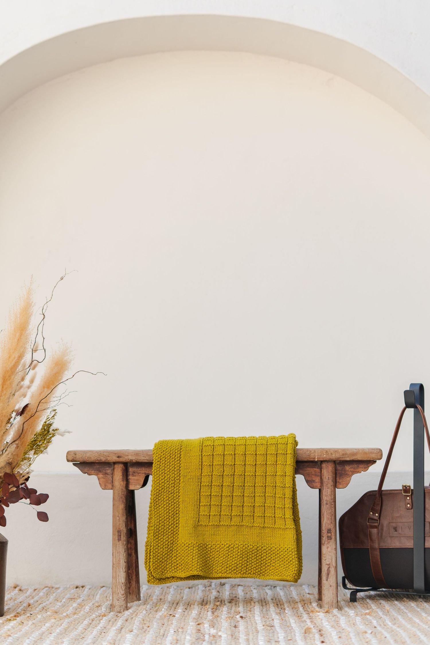 Rustic wooden bench with mustard yellow knit throw under white arch, styled with dried pampas grass and leather bag.