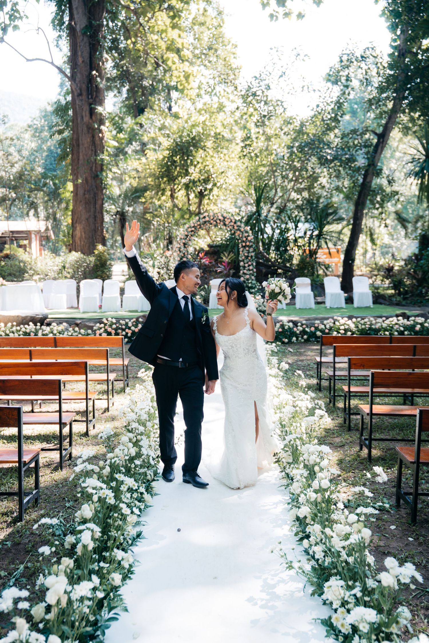 Newlywed couple walks down floral aisle at outdoor garden wedding ceremony, groom raising hand in celebration.