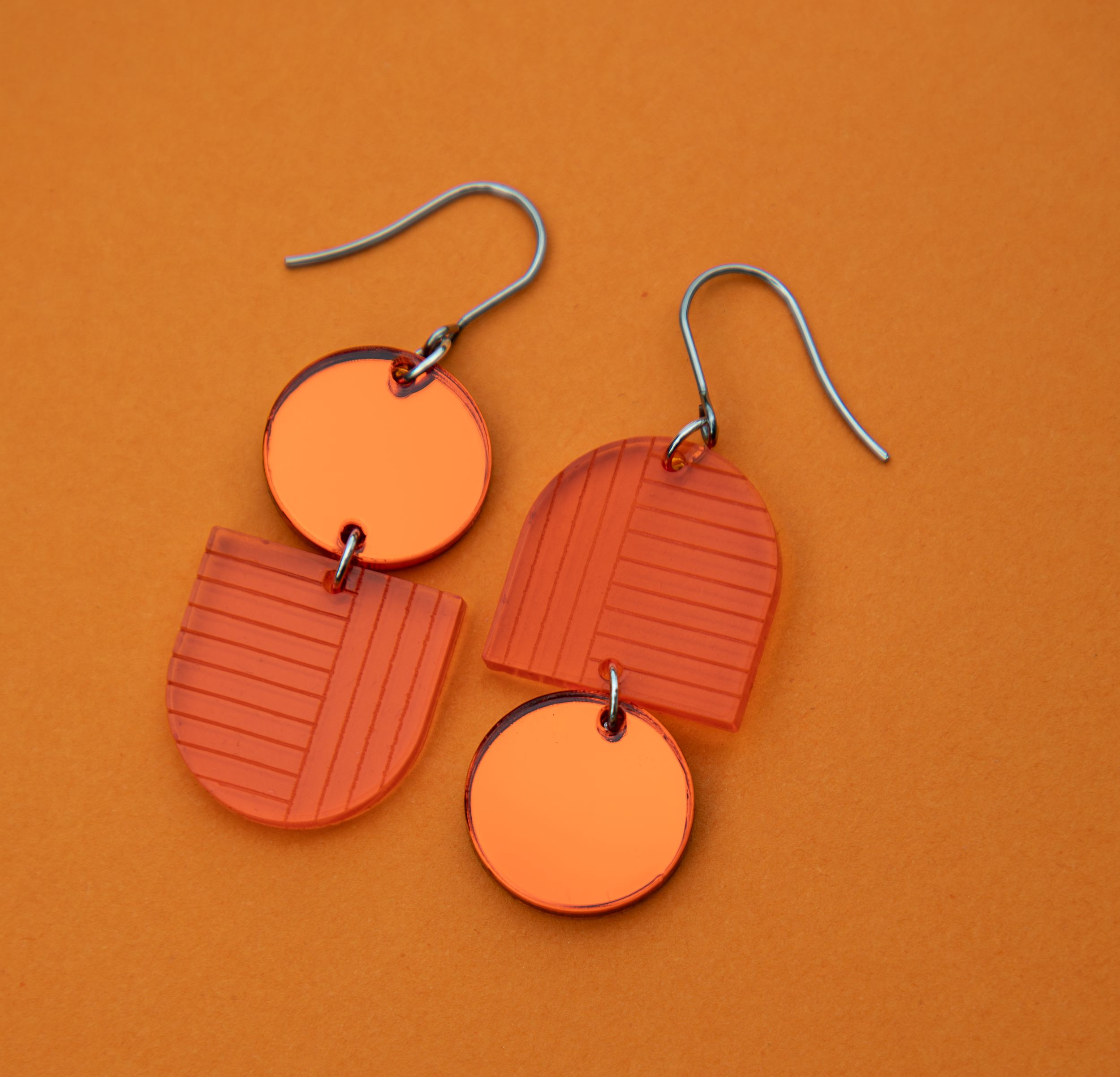 Pair of orange geometric two-piece dangle earrings with circle and arch shapes on an orange background.