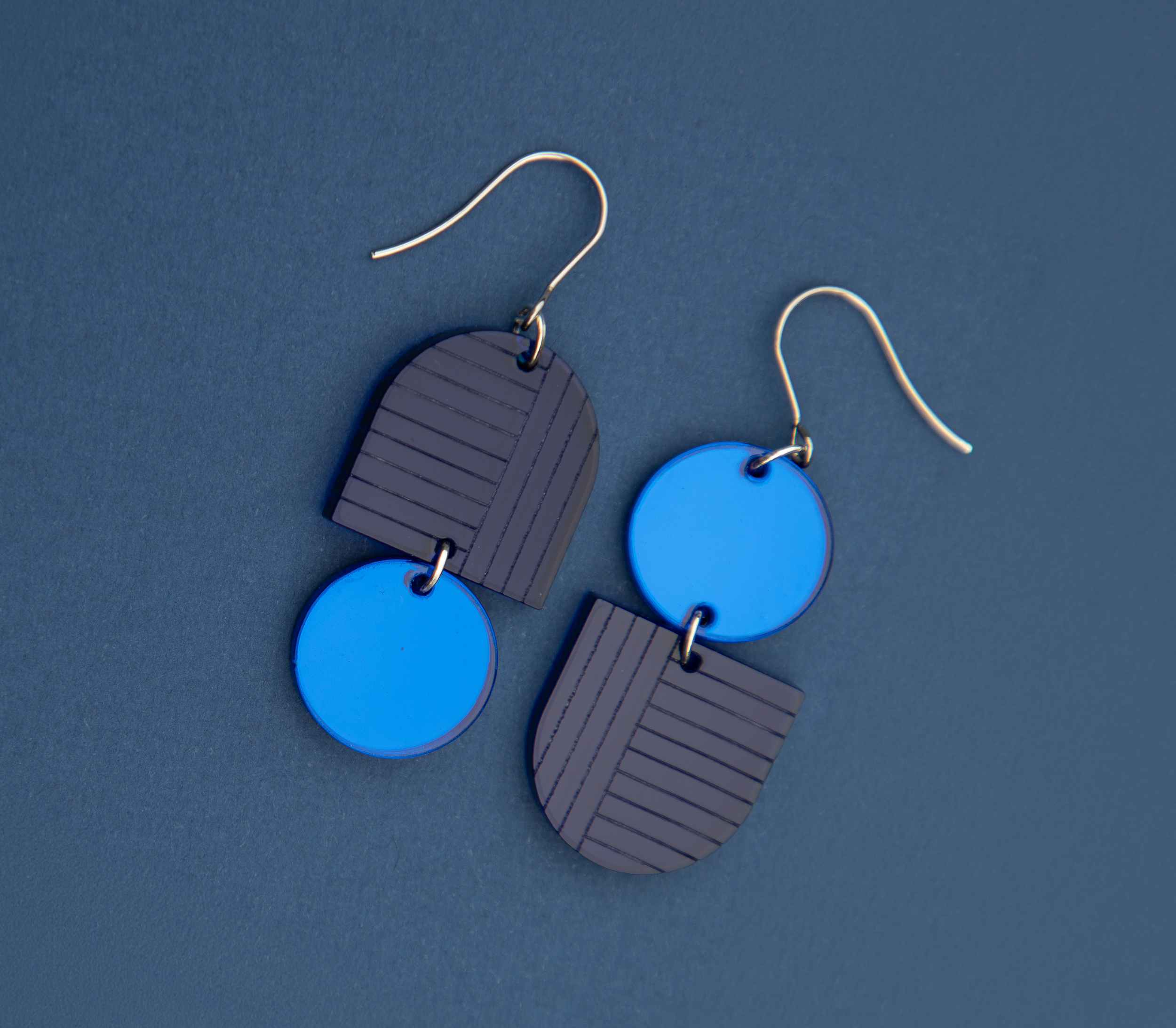 Pair of navy blue and bright blue two-tone geometric arch and circle dangle earrings on a dark blue surface.