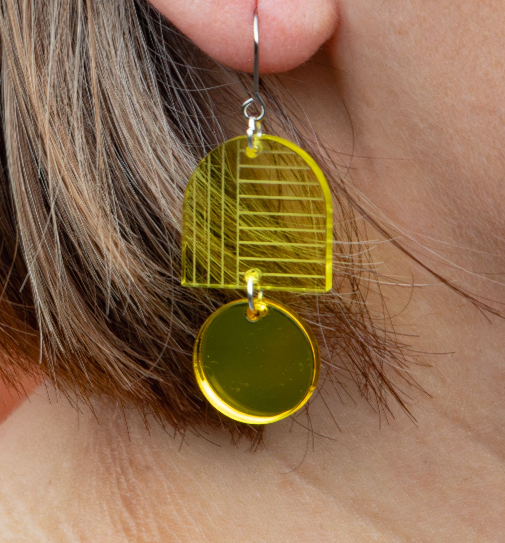 Close-up of a yellow geometric arch and circle dangle earring made from transparent acrylic worn on an ear.