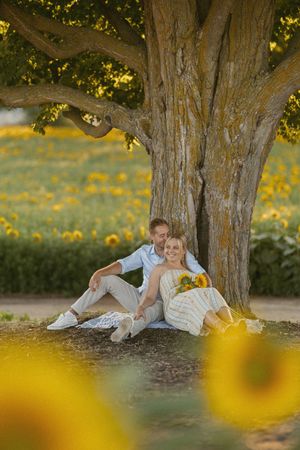 Couple sitting under large tree in sunflower field, woman holding yellow bouquet, enjoying a romantic outdoor moment.