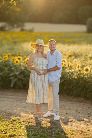 Pregnant couple posing in sunflower field at golden hour, woman in striped dress and hat, man in blue shirt.