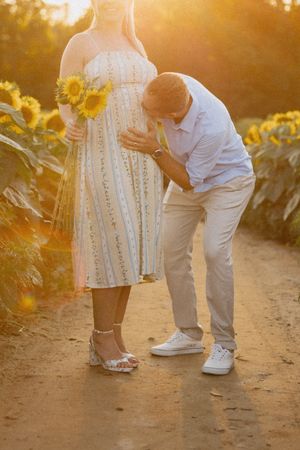 Couple in sunflower field at golden hour, man kissing pregnant woman's belly, she holds sunflowers.