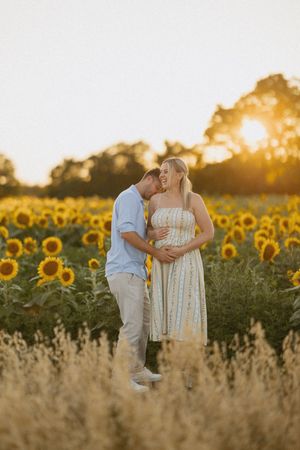 Couple embracing in a sunflower field at golden hour, woman in floral dress with baby bump, man in blue shirt.