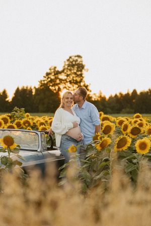 Couple sharing a tender moment in sunflower field at sunset, pregnant woman leaning on vintage car.