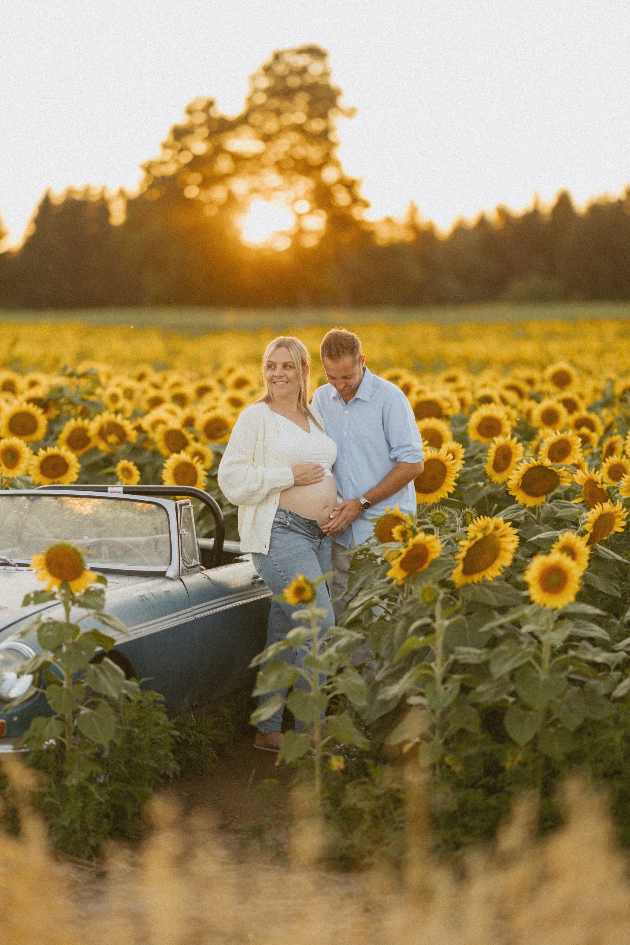 Pregnant couple standing in a sunflower field at golden hour beside a vintage convertible car.