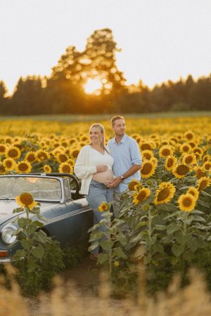 Pregnant couple embracing by vintage convertible in sunflower field at sunset, golden light.