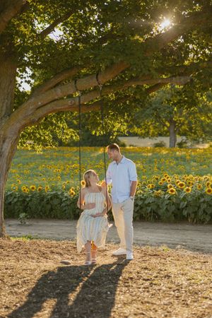 Couple standing by a tree swing in front of a sunflower field during golden hour.