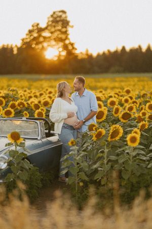 Couple gazing at each other beside classic car in sunflower field during golden hour sunset.