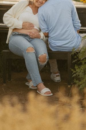 Pregnant woman in ripped jeans and white sandals sits with partner who tenderly touches her baby bump on a bench outdoors.