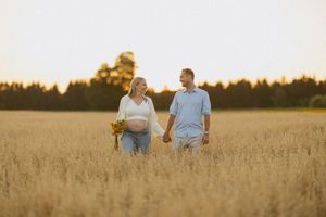 Pregnant couple holding hands in golden wheat field at sunset, woman cradling baby bump with wildflowers.
