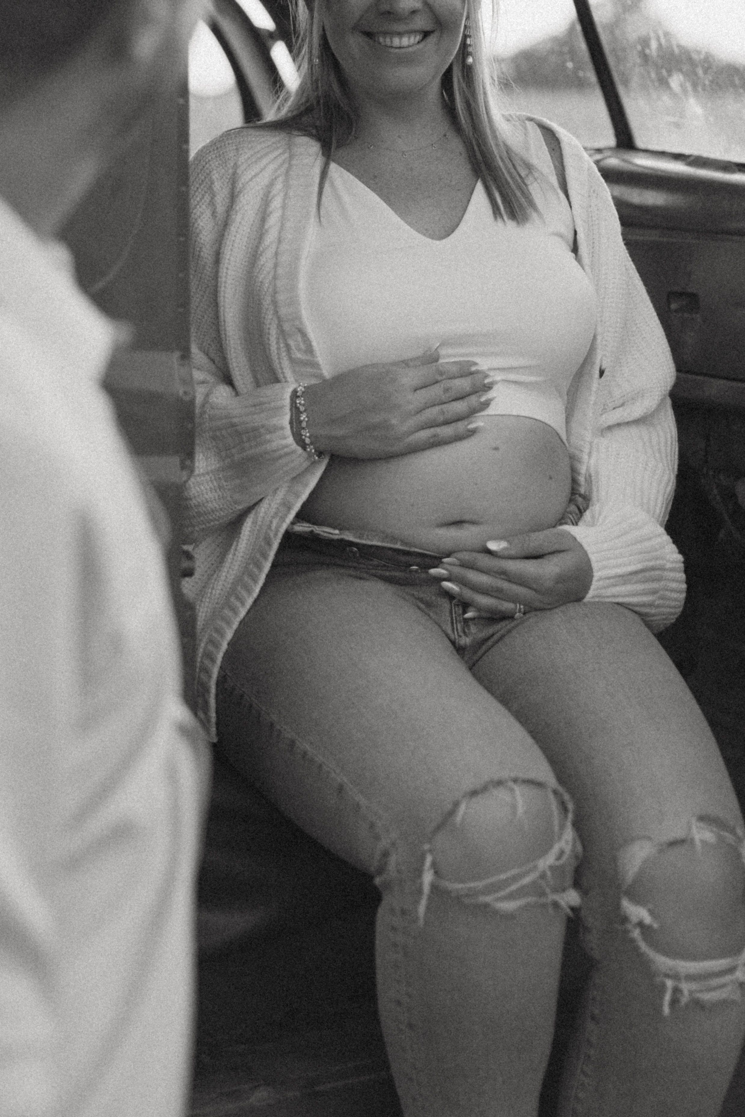 Black and white photo of a smiling pregnant woman sitting in a vintage car, cradling her baby bump.