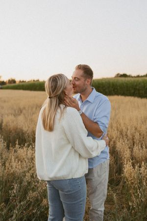 Couple sharing a romantic moment in a golden field at sunset, woman in white sweater and man in blue shirt embracing.
