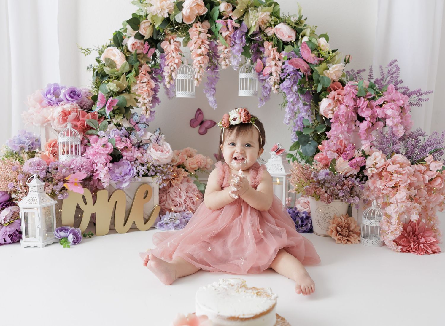 Baby girl in pink dress celebrates first birthday with floral arch backdrop and cake smash setup.