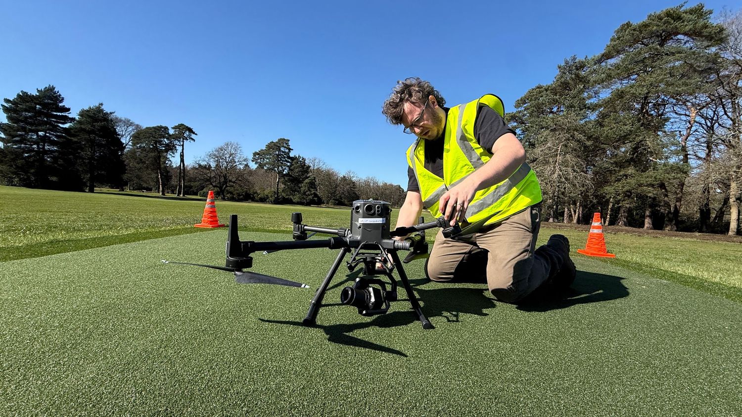 Technician in hi-vis vest preparing a professional drone on green turf outdoors on a sunny day.