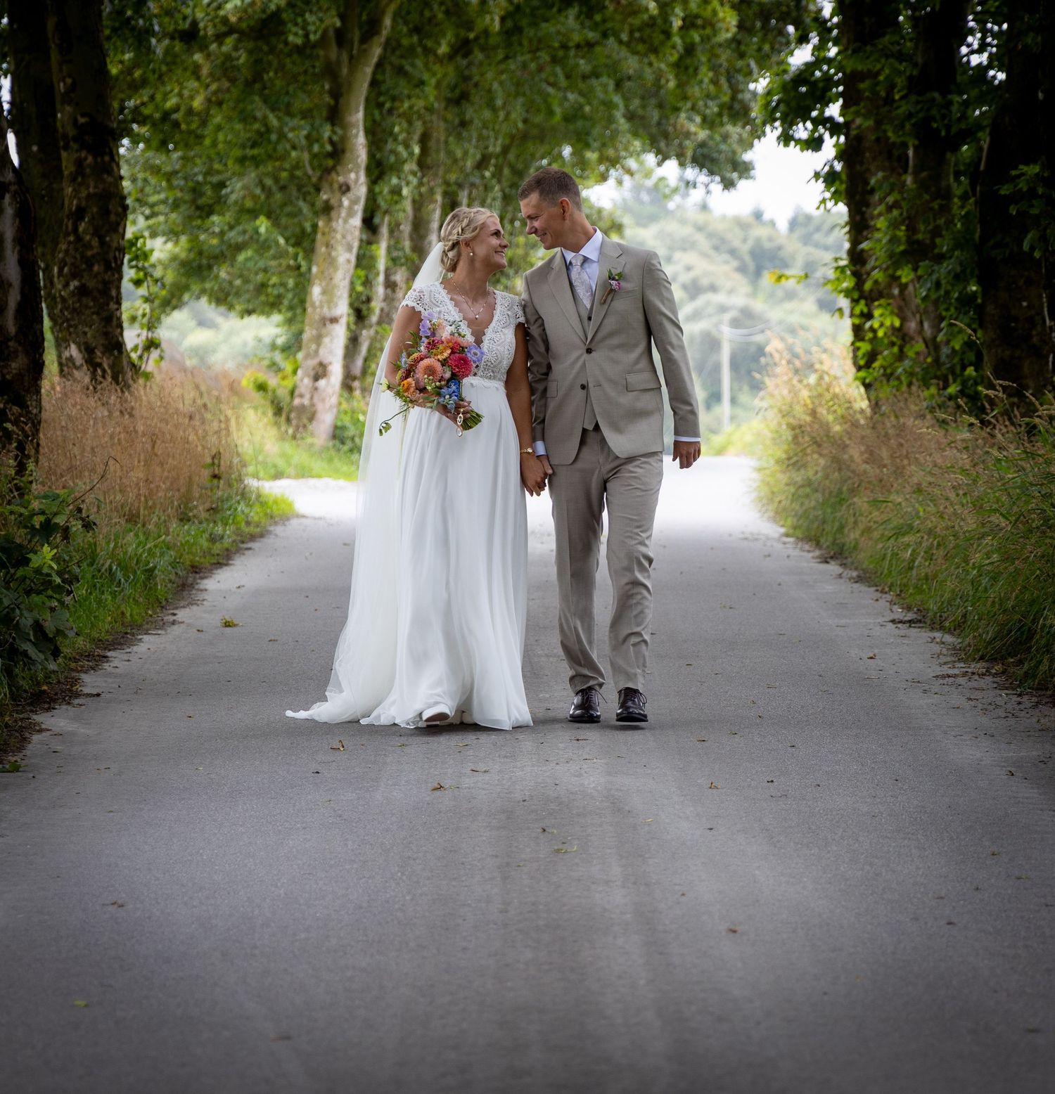 Bride and groom walking hand in hand down a tree-lined road, bride in white gown holding colorful bouquet.