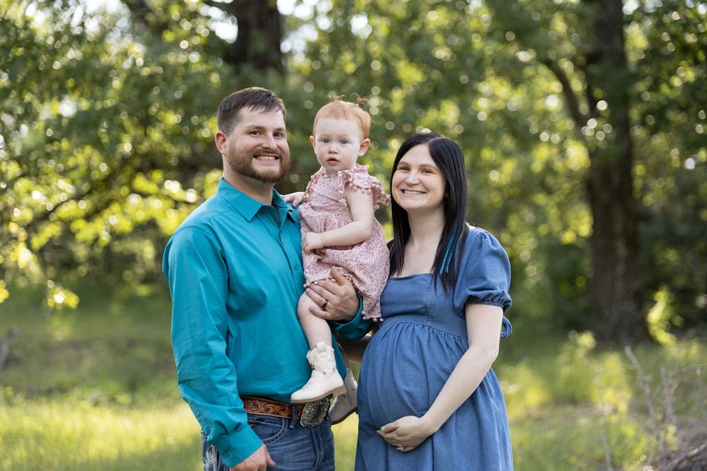 Smiling family with pregnant mother, father in teal shirt, and baby girl posing outdoors in a lush green park setting.
