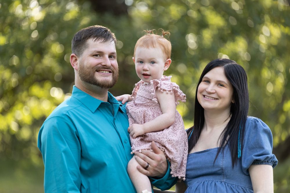Happy young family with parents smiling and holding their baby girl outdoors in a lush green park setting.