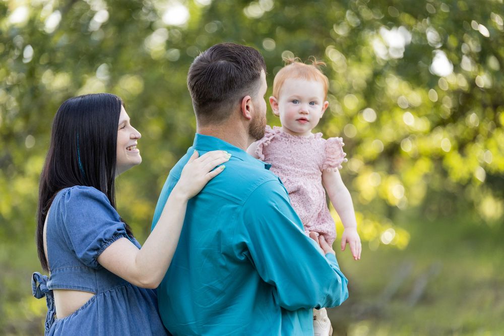 A young family with a redhead baby girl outdoors, parents smiling in a lush green park setting.