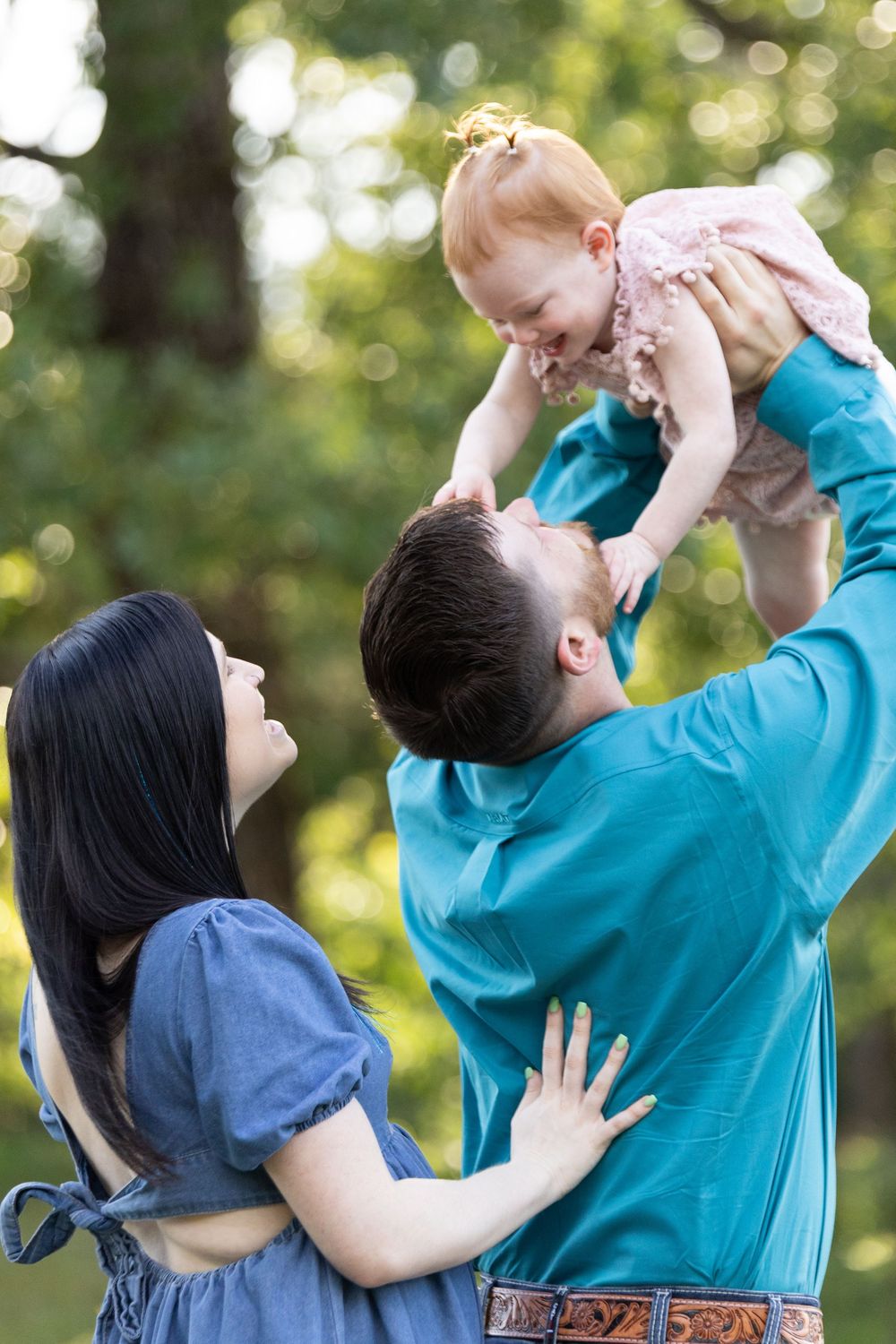A happy family with a father lifting a laughing baby girl in a pink dress outdoors, while the mother smiles beside them.