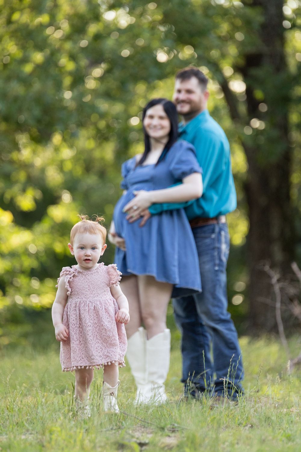 A toddler girl in a pink dress stands in a grassy field as her pregnant mother and father embrace behind her.