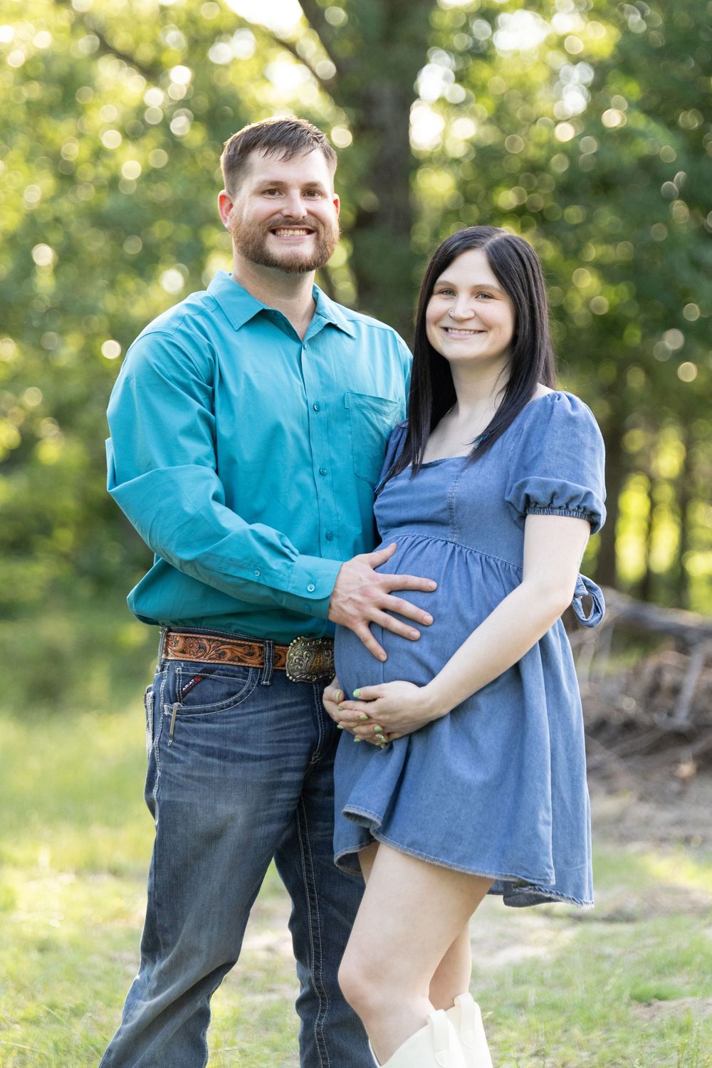 A smiling couple poses outdoors, the woman in a blue dress showing a baby bump, the man in a teal shirt embracing her.