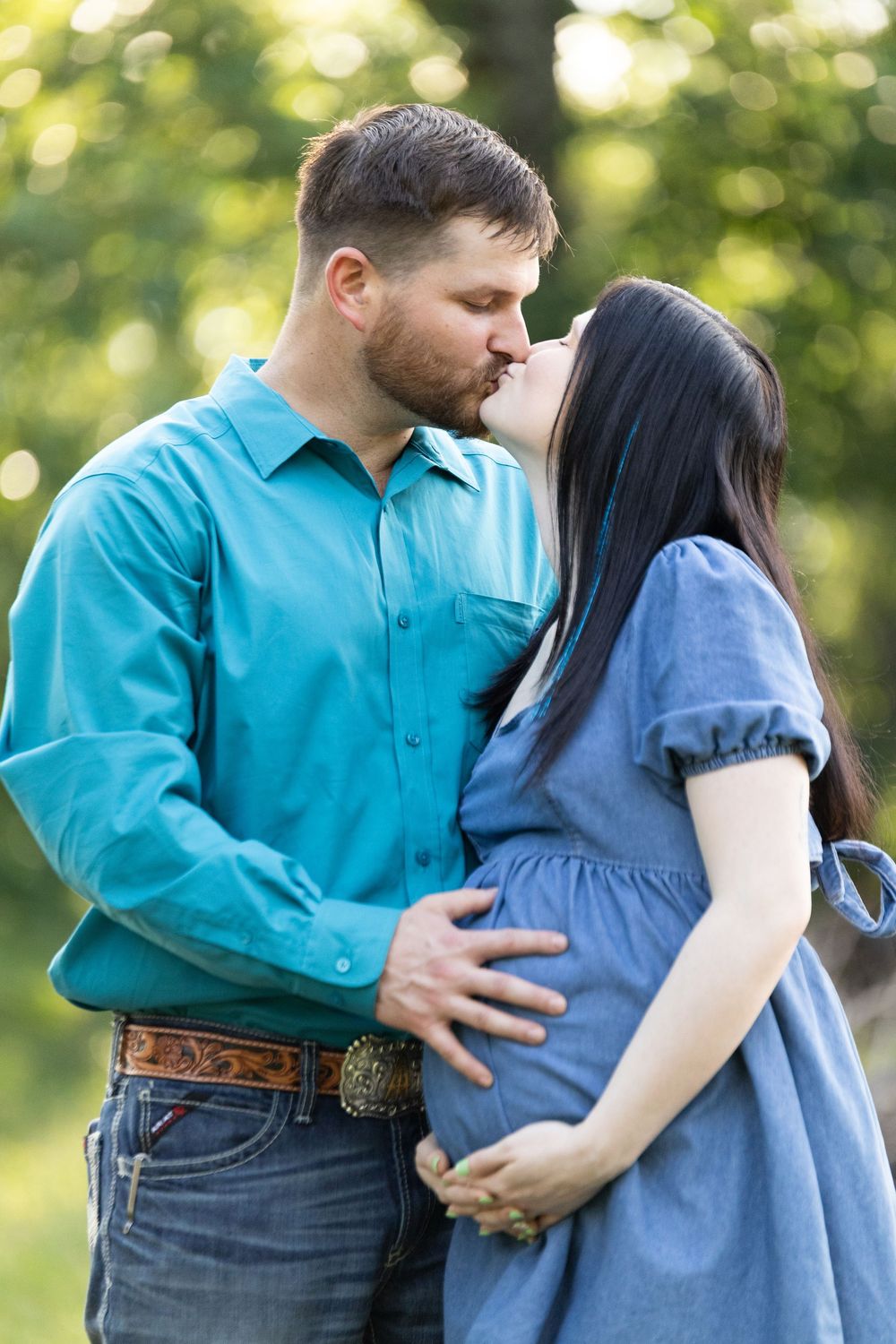 A couple shares a tender kiss outdoors, with the man cradling his pregnant partners baby bump in a lush green setting.