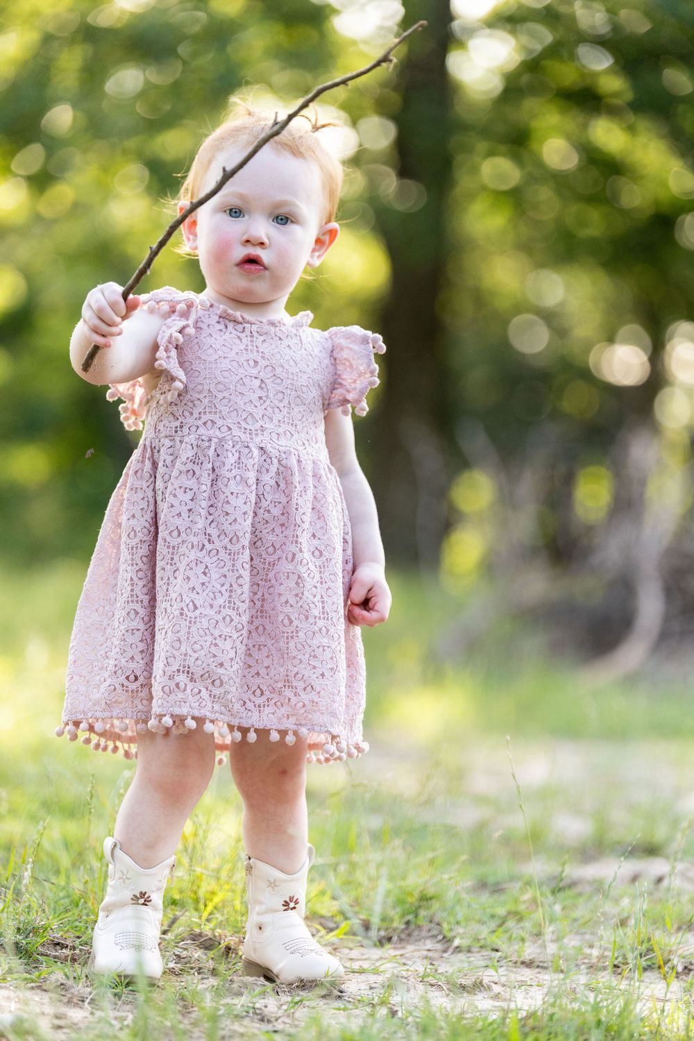 Adorable toddler girl in pink lace dress holding a stick outdoors in a sunny green park setting.