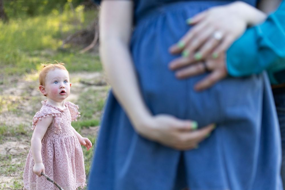 Pregnant woman in blue dress cradles baby bump outdoors while red-haired toddler girl in pink dress stands nearby.