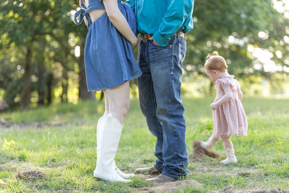 A couple stands close together outdoors while a toddler girl in a pink dress plays nearby on grass.