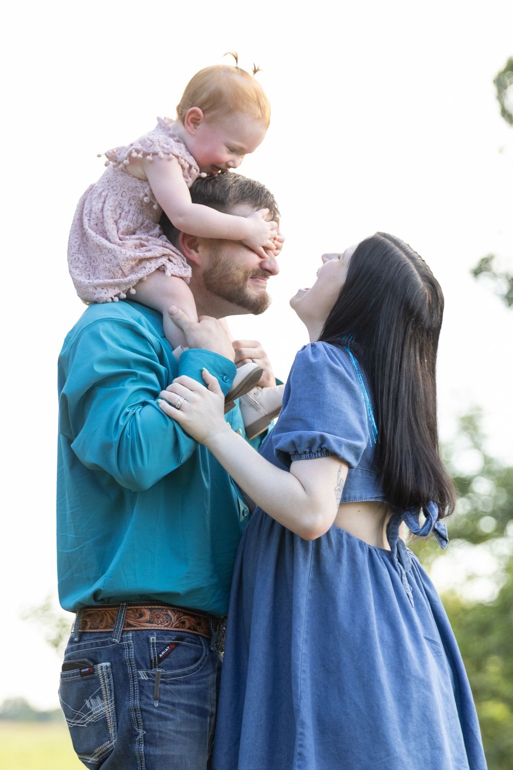 A happy family outdoors, father holding baby on shoulders while mother smiles lovingly at them in a sunny park.