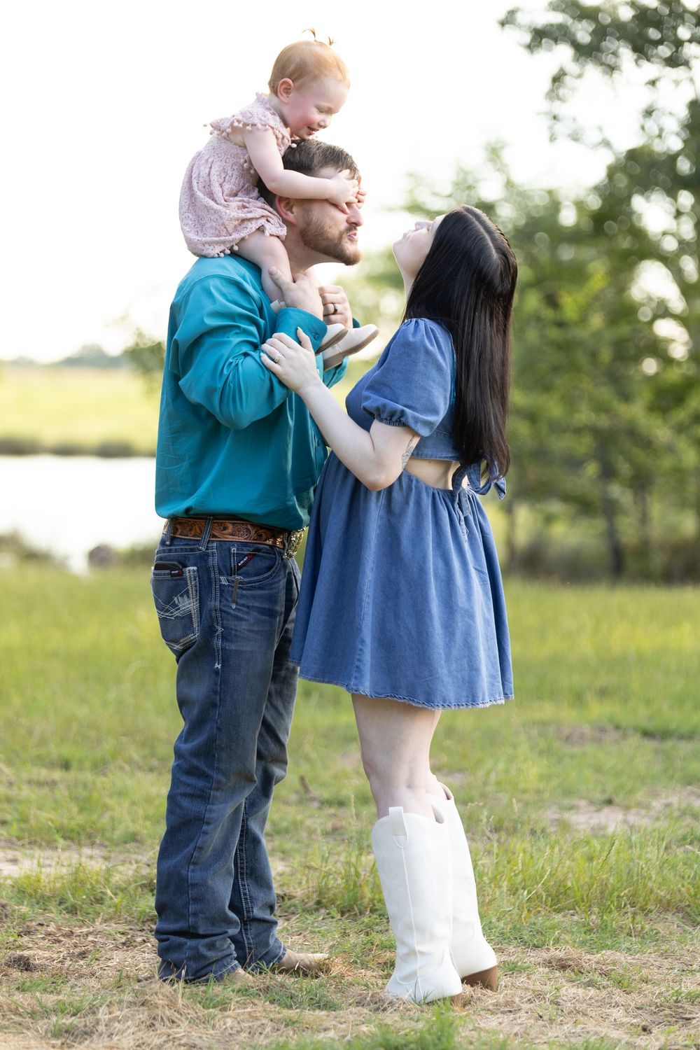 A happy young family outdoors, father holding baby girl on shoulders while mother in blue dress and white boots smiles up at them.