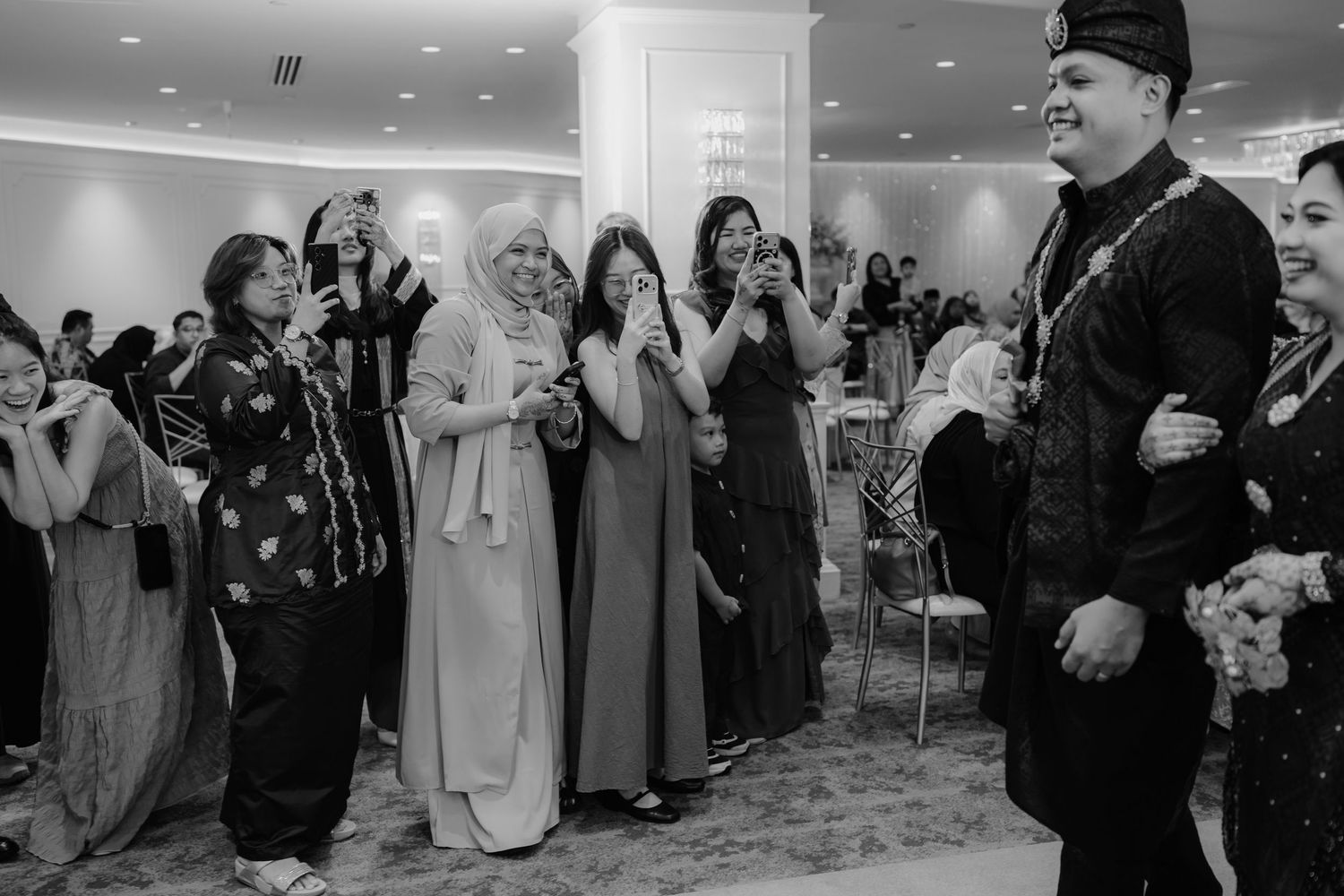Traditional Malay wedding ceremony with couple seated on stage, performers in green, floral decor, and waterfront backdrop.
