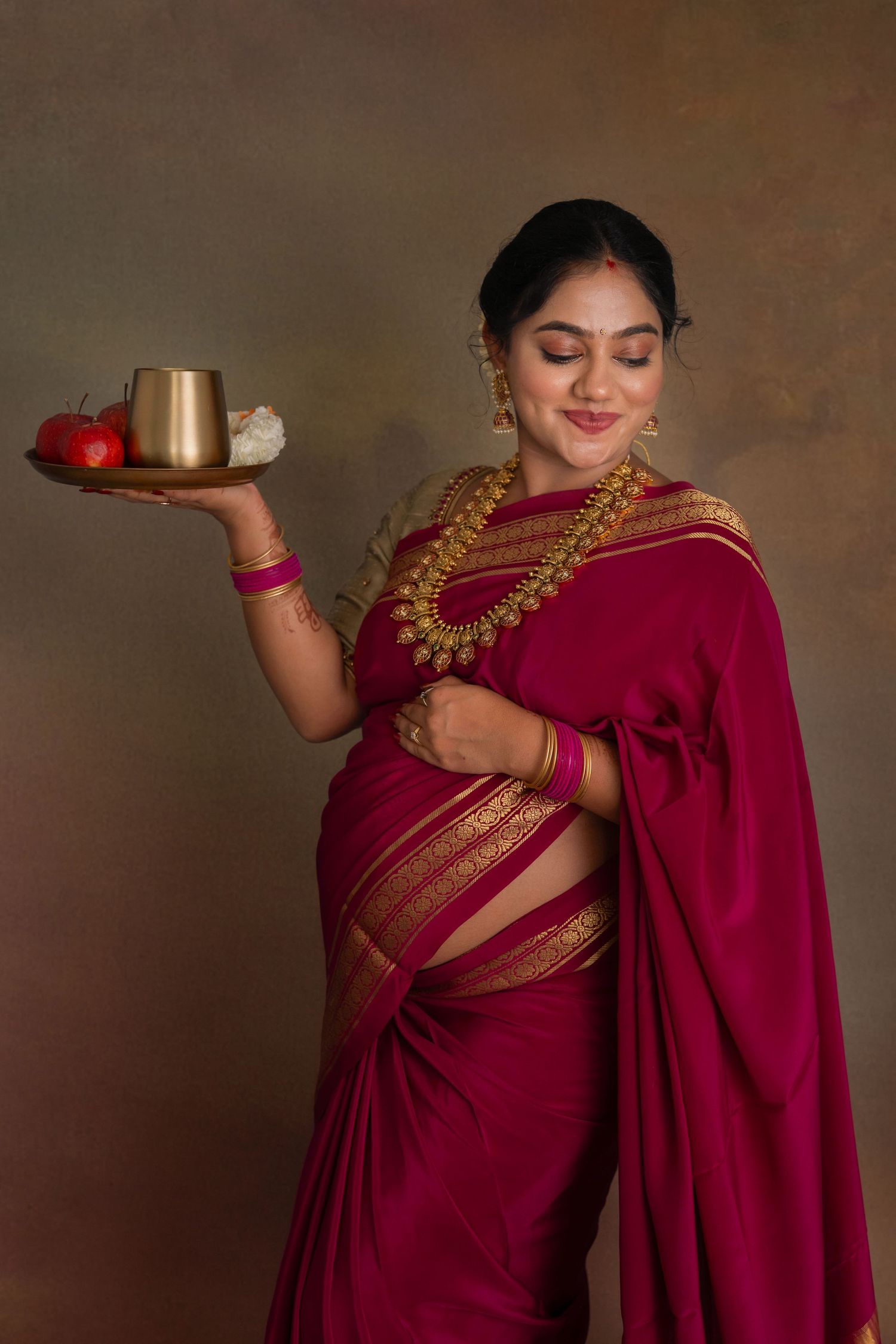 Expecting mother in a magenta silk saree holding a ceremonial thali with fruits and flowers during a Seemantham-style maternity session
