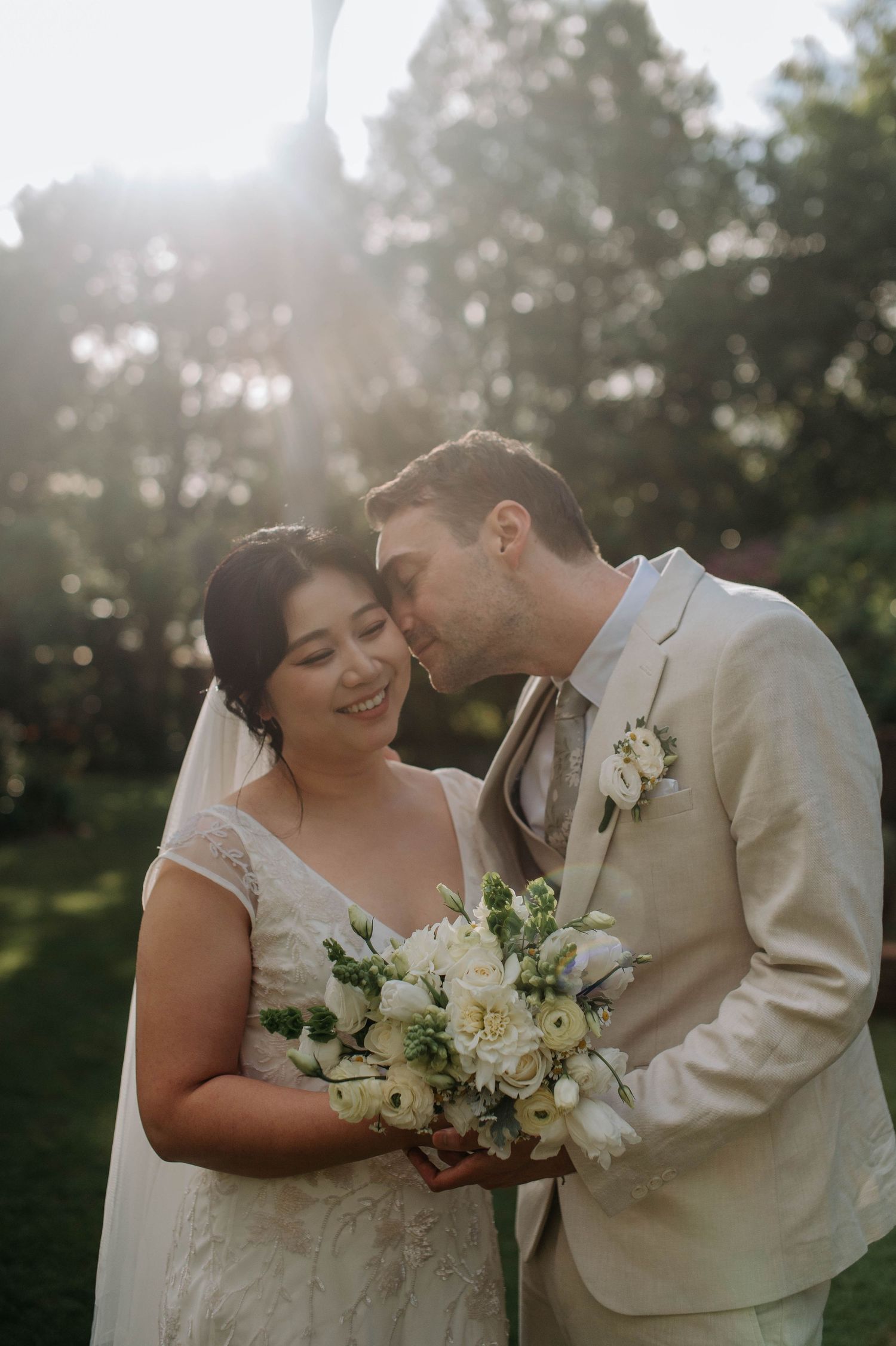 A bride and groom share a tender moment outdoors, with golden sunlight glowing behind them as she holds a white bouquet.