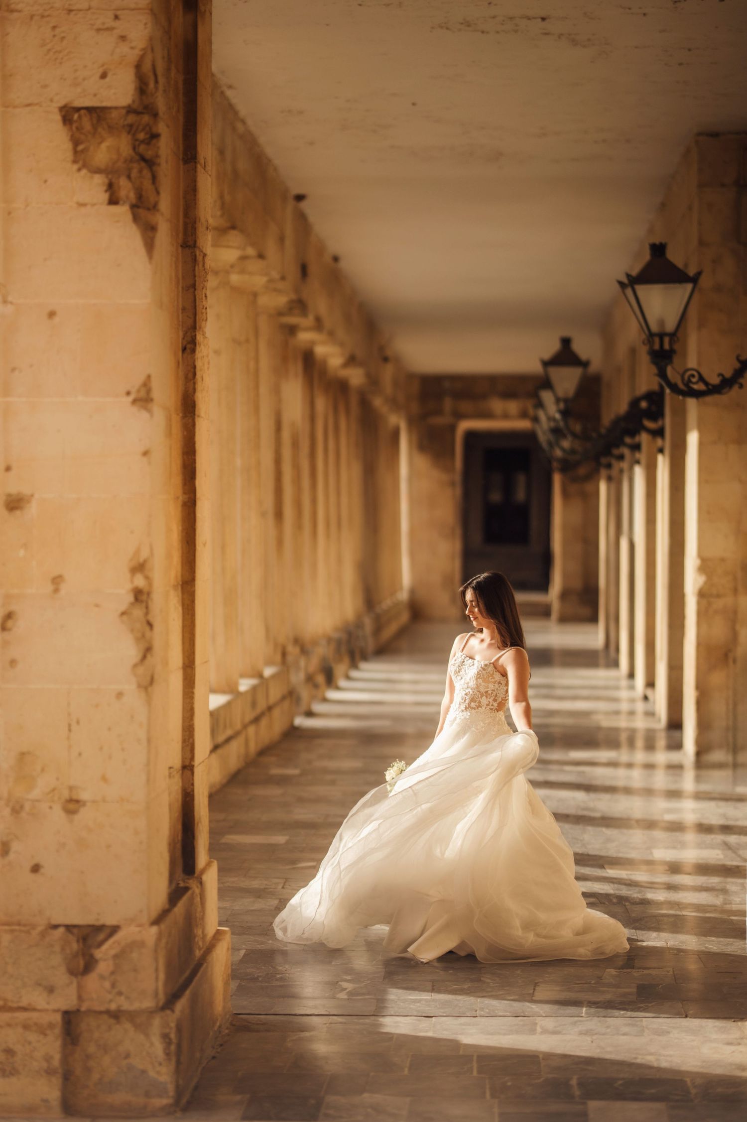 Bride in flowing white gown walking through elegant sunlit colonnade with marble columns and vintage lanterns.