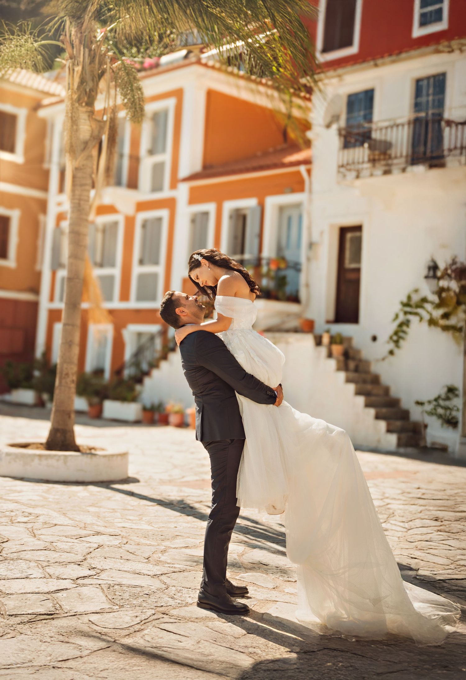 Groom lifts bride in white gown outside colorful Mediterranean building on cobblestone plaza with palm tree.